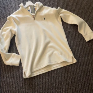 Vit half zip tröja från Polo Ralph Lauren - Snygg vit långärmad tröja från Polo Ralph Lauren med klassisk half zip-dragkedja och broderad logga på bröstet. Tröjan är i mjuk bomull och har en stilren, normal passform. Perfekt till lager-på-lager och enkel att matcha med jeans eller chinos. Helt oanvänd