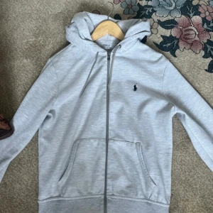 Ljusgrå hoodie från Polo Ralph Lauren - Säljer en stilren ljusgrå hoodie från Polo Ralph Lauren med dragkedja och klassisk broderad logga på bröstet. Tröjan har huva med snörning och två fickor framtill. Perfekt för en chill och avslappnad look.