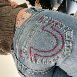 True Religion ljusblå bootcut jeans - Säljer ett par ljusblå jeans från True Religion med ikoniska röda sömmar på bakfickorna och klassisk patch i midjan. Jeansen har bootcut-modell och normal passform, tillverkade i jeansmaterial. Perfekta för dig som gillar en avslappnad men snygg look. Använda fåtal gånger