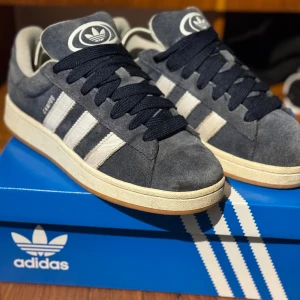 Adidas Campus 00s mörkblå sneakers - Adidas Campus 00s sneakers i mörkblå mocka med vita klassiska stripes och beige sula. Skorna har svart snörning, rund tå och gummisula med fiskbensmönster. Stor Adidas-logga på plösen och retrovibbar. Perfekta för dig som gillar streetstyle och tidlös design. Kvitto och extra snören finns  sparsamt andvända