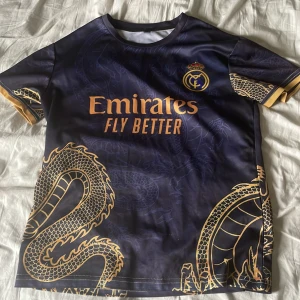 Real Madrid Ronaldo 7 draktröja - Snygg fotbollströja från Real Madrid med Ronaldo 7 på ryggen. Tröjan är mörkblå med guldiga drakdetaljer och tryck, samt klubbmärke och Emirates-logga på bröstet. Tillverkad i lätt och sval polyester, perfekt för match eller träning.