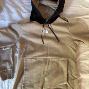Beige hoodie från The North Face - Snygg beige hoodie från The North Face med svart huva och vita logotyper fram och bak. Tröjan har långa ärmar, ribbade muddar och en stor magficka. Perfekt för chill dagar och streetwear-stil.