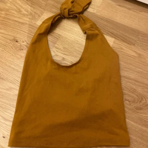 Brun ärmlös halterneck-linnetopp - Säljer en brun ärmlös halterneck-linnetopp med knyt i nacken. Toppen har en enkel och clean design i mjukt bomullsmaterial, perfekt för varma dagar. Snyggt djup ringning framtill och croppad passform som ger en trendig look.