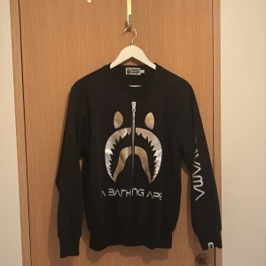 Svart crewneck från A Bathing Ape - Svart crewneck-tröja från A Bathing Ape med stort metallic-silver och guld tryck framtill och texten 'SORAYAMA' i silver på ena ärmen. Klassisk rund hals och ribbade muddar. Perfekt statement piece för streetwear-stilen.