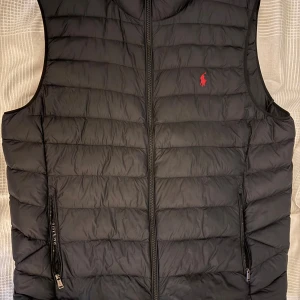 Svart dunväst från Ralph Lauren - Snygg svart dunväst från Ralph Lauren med röd broderad logga på bröstet. Västen har quiltad design, två fickor med dragkedja och hel dragkedja framtill. Perfekt för lager-på-lager och stilren look.