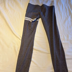 Grå leggings med vita detaljer - Snygga grå leggings med bred ljusgrå midja och vita ränder samt tryck på ena benet. Perfekta för träning eller chill, med stretchigt material som sitter skönt och följer kroppen. Sportig look med coola detaljer som sticker ut.