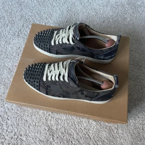 Christian Louboutin junior spikes  - Snygga sneakers från Christian Louboutin med kamouflagemönster i mocka och ikoniska röda sulor. Skorna har vita snören och coola svarta nitar på tån för en edgy vibe. Perfekta för dig som vill sticka ut med exklusiva detaljer. Box och extra snören ingår✌️, storlek 41 passar 41,5 också