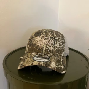 Stüssy x New Era camo keps - Säljer en Stüssy x New Era 9TWENTY keps med justerbar passform. Kepsen har camouflage-mönster i brunt, beige och vitt samt broderad Stüssy-logga framtill. Baksidan är i mesh för extra ventilation. Perfekt för dig som gillar streetwear och snygga accessoarer.
