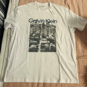 Vit Calvin Klein t-shirt med citytryck - Vit t-shirt från Calvin Klein i 100% bomull med ett stort svartvitt citymotiv på bröstet och tydlig Calvin Klein-logga ovanför. Klassisk rund hals och korta ärmar. Perfekt för dig som gillar stilrena plagg med en urban touch.