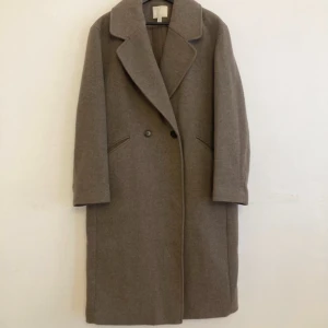 Beige ullkappa med bred krage - Stilren beige kappa  med bred krage och dubbelknäppning framtill. Lång modell med raka ärmar och diskreta fickor i sidorna. Perfekt för dig som gillar en klassisk och clean look under höst och vinter.