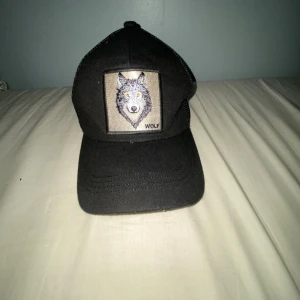Svart keps med vargmärke och mesh - Svart keps med broderad varg på framsidan och texten 'WOLF' under. Framsidan är i tyg och baksidan har mesh för extra ventilation. Klassisk böjd skärm och justerbar passform. Perfekt för dig som gillar djurmotiv och streetstyle.