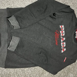 Mörkgrå sweatshirt från Prada - Snygg mörkgrå sweatshirt från Prada med broderad logga i vitt och rött på bröstet samt en mindre röd Prada-detalj på ärmen. Tröjan har rund hals, ribbade muddar och är tillverkad i mjuk bomullsmix. Perfekt för en chill och trendig look.