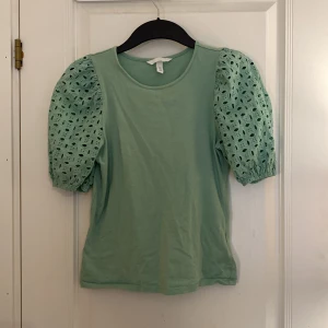 Mintgrön topp med spets puffärmar från H&M - Superfin mintgrön topp från H&M med korta puffärmar i broderad bomull. Ärmarna har elastiska muddar och coola håldetaljer som ger en unik look. Perfekt för dig som vill sticka ut med en söt och trendig stil. Perfekt tröja till våren för dig som vill ha en unik tröja med en vågig färg och fina detaljer.