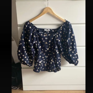 Blommig offshoulder blus navy - Supersöt blommig blus i mörkblått med vita och gula små blommor. Offshoulder-modell med vida trekvartsärmar och knappar framtill. Perfekt för en chill och trendig look.