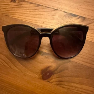 Svarta solglasögon från H&M - Snygga svarta solglasögon från H&M med rundad, något kattig form. Bågarna är i blank plast och glasen är mörkt tonade. Perfekta för att ge din look en cool vibe under soliga dagar.