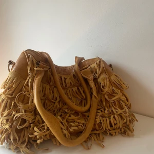 Beige axelväska med fransar i skinn - Cool beige axelväska i skinn med massor av fransar över hela väskan. Väskan har två rejäla handtag och detaljer i silverfärgad metall. Perfekt för dig som vill sticka ut med en bohemisk och trendig look.