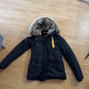 Svart Parajumpers parkajacka med päls - Svart parkajacka från Parajumpers med stor huva fodrad med mjuk pälsimitation och pälsbräm. Jackan har flera fickor, gul detaljrem och dragkedja framtill. Perfekt för kalla dagar och har en klassisk, stilren look. Dragkedjan har gått sönder i kanten men inget som påverkar stängningen, se bild. Jackan säljs i befintligt pris, om fler bilder eller funderingar så skriv.