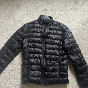 Svart pufferjacka från EA7 Armani - Snygg svart pufferjacka från EA7 Emporio Armani med quiltad design och diskret logga på bröstet. Jackan har dragkedja framtill, hög krage och långärmad passform. Perfekt för kyliga dagar och ger en clean, modern look.
