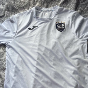 Vit Joma CS Academy fotbollströja S - Barcelona tröja använd 1 gång säljs pga att den är för stor. Vit fotbollströja från Joma i storlek S med CS Academy Barcelona-logga på bröstet och fyra guldstjärnor ovanför. Tröjan är tillverkad i ett lätt material för träning.  svår att få. Kontakta innan köp 
