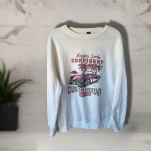 Vit sweatshirt med biltryck  - Vit sweatshirt  med coolt tryck av rosa bil, palmer och texten 'Happy Smile Confident Custom Motors'. Tröjan har rund hals, lång ärm och är gjord i mjuk polyesterblandning. Perfekt för dig som gillar retrovibes och streetstyle.