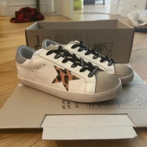 Golden Goose sneakers med glitter och leopard - Snygga vita Golden Goose sneakers med svart text på snörningen, silverglittrig häl och beige mockadetaljer på tån. Sidan har en stjärna i leopardmönster som ger extra edge. Perfekta för dig som vill sticka ut med coola detaljer och streetstyle-vibe.