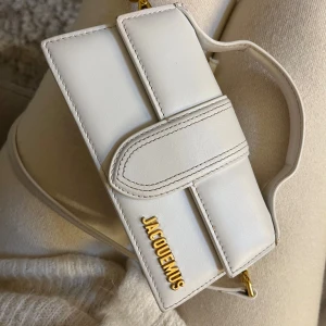 Jacquemus Le Chiquito Bag 👜 - Säljer den populära Le Chiquito-väskan från Jacquemus i vitt läder. Klassisk modell med topphandtag, guldfärgad logotyp och avtagbar axelrem som gör den enkel att bära både i handen eller crossbody. Handgjord i Italien.  	•	Färg: Vit 	•	Material: Läder 	•	Detaljer: Guldfärgad logotyp 	•	Kategori: Mini-väska / crossbody