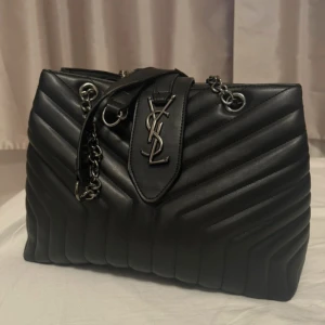 Svart quiltad axelväska  - Snygg svart axelväska med quiltad design och silverfärgade kedjedetaljer. Väskan har det ikoniska YSL-emblemet framtill och är tillverkad i skinn. Perfekt för dig som vill ha en lyxig och stilren accessoar.