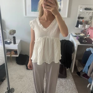 Vit volangblus med v-ringning - Säljer en söt vit blus med v-ringning och volangdetaljer vid ärmslut och midja. Blusen är ärmlös med lätt rynkad struktur och har en luftig, flowy passform. Perfekt för dig som gillar romantisk och avslappnad stil.