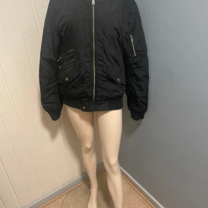 Svart bomberjacka med dragkedjor - Cool svart bomberjacka med flera fickor och silvriga dragkedjor. Jackan har ribbade muddar vid ärmslut och midja, samt klassisk bomberpassform. Perfekt för dig som gillar streetstyle och vill ha en jacka med attityd storlek är S men passar M också 