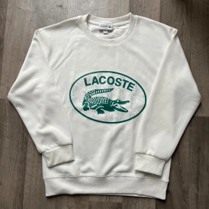 Vit sweatshirt från Lacoste med logga - Vit sweatshirt från Lacoste i regular fit med grön krokodil-logga och text på bröstet. Tröjan har rund hals, långa ärmar och ribbade muddar. Tillverkad i mjuk bomullsmix som känns skön mot huden. Perfekt för dig som gillar klassisk och clean stil.
