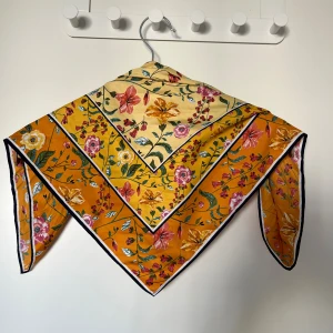Blommig gul och orange scarf - Snygg scarf i kvadratisk modell med blommigt mönster i gult och orange. Tunt och glansigt material som känns lyxigt och lätt att styla runt halsen eller i håret. Sparsamt använd, 73 cm i diameter