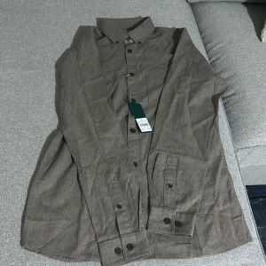 Brun långärmad skjorta med knappar - Snygg brun skjorta från H&M i bomull med klassisk krage och svarta knappar framtill. Skjortan har lång ärm med manschett och en bröstficka. Perfekt för dig som gillar en clean och stilren look.