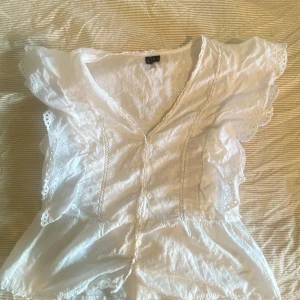 Vit blus med volang och spetsdetaljer - Superfin vit blus från vero Moda med v-ringning, knappar framtill och volangärmar. Blusen har broderade spetsdetaljer och små prickar i tyget. Perfekt för en somrig look med romantiska vibes.