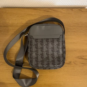 LACOSTE Axelväska - Mörkgrå Lacoste axelväska. Sjukt bra material och hållbarhet, endast använd 1 gång och är i jättebra skick.                                                                     - Pris kan diskuteras