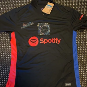 FC Barcelona svart matchtröja Nike M - Snygg svart FC Barcelona fotbollströja från Nike med röda och blå detaljer på sidorna. Tröjan har klubbmärke och Nike-logga på bröstet, samt Spotify som sponsor framtill. Tillverkad i lätt och ventilerande material, perfekt för match eller träning.