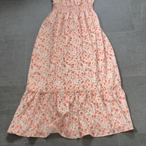 Blommig långklänning med volangkant - Söt långklänning i ljusbeige med små röda och gröna blommor. Klänningen har smock upptill och en bred volang längst ner för extra flow. Perfekt för varma dagar och har en romantisk vibe. Materialet känns lätt och luftigt.