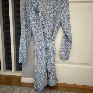 Blåvit omlottklänning med volang - Supersöt blåvit omlottklänning med blommigt mönster och volangdetaljer längs kanten. Klänningen har långa ärmar och knyts i midjan för en snygg passform. Perfekt för vår och sommar när du vill känna dig extra fin.