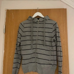 Randig kofta  - Grå hoodie med svarta ränder och hel dragkedja framtill. Tröjan har huva med snören och två fickor med dragkedja.