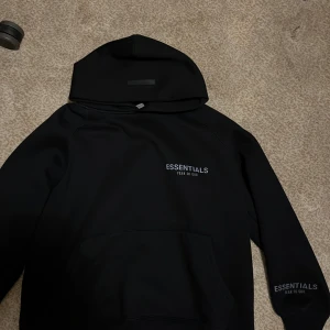 Svart Essentials Fear of God hoodie - Svart hoodie från Essentials Fear of God med tryck på bröstet och ärmen. Klassisk känguruficka framtill och stor huva. Oversized passform och mjukt material som känns riktigt skönt. Perfekt för dig som gillar streetwear och enkel stil.