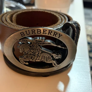 Brunt skärp från Burberry med stor spänne - Säljer ett brunt skärp från Burberry med ett stort, ovalt metallspänne som har en detaljerad riddare och texten 'Prorsum'. Skärpet är gjort i skinn och har vita kontrastsömmar längs kanterna. Perfekt för att lyfta vilken outfit som helst. 