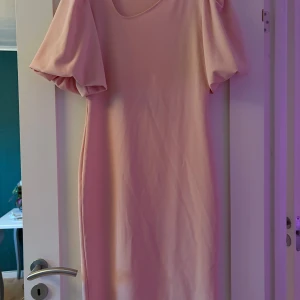 Ljusrosa klänning med puffärm - Superfin ljusrosa klänning med puffiga korta ärmar och assymetrisk rund halsringning. Klänningen har en rak passform och är sydd i ett mjukt, lätt material som ger ett snyggt fall. Perfekt för dig som gillar en enkel men söt stil.