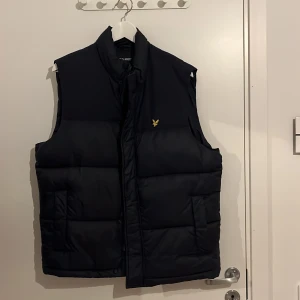 Svart dunväst från Lyle & Scott - Snygg svart dunväst från Lyle & Scott med klassisk gul logga på bröstet. Västen är quiltad och har hög krage samt två fickor framtill. Perfekt för lager-på-lager och enkel att matcha med olika outfits.
