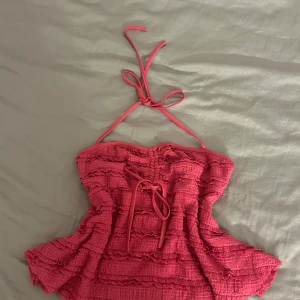 Rosa volangig halterneck tubtopp - Supersöt rosa tubtopp med halterneck och volanger i flera lager. Toppen har knytband både i nacken och framtill, vilket ger en extra detalj. Materialet är lätt och strukturerat, perfekt för varma dagar eller när du vill sticka ut lite extra.