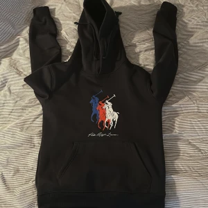Svart hoodie från Polo Ralph Lauren - Svart hoodie från Polo Ralph Lauren med stor färgglad polospelare-tryck i blått, rött och vitt på bröstet🔥. Klassisk känguruficka, huva med snörning och mjukt material. Inga defekter eller något, tvär bra passfor💯. Säljer för jag har för mycket raffe plagg😬Nypris 2300kr. Skriv om några frågor!