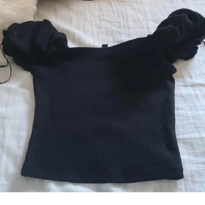 Svart croppad topp med puffärmar Divided - Säljer en svart croppad topp från Divided med ribbad struktur och stora puffärmar. Toppen är axelbandslös och sitter tight över kroppen, perfekt för en trendig look. Materialet är mjukt och stretchigt, vilket gör den bekväm att bära.