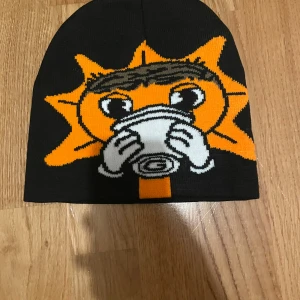 Svart mössa med sol och kaffemotiv - Cool svart mössa med ett stort broderat motiv av en tecknad sol som dricker kaffe. Motivet har starka färger i orange, vitt och brunt, vilket gör mössan till en riktig blickfångare. Perfekt för dig som vill sticka ut och gillar roliga accessoarer.