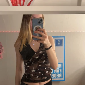 Y2k Camo omlott-topp med stjärnor - Säljer en omlott-topp med camouflage-mönster i bruna och svarta toner, dekorerad med vita stjärnor. Toppen har svarta detaljer, ringar vid axelbanden och knytning i nacken. Sitter tight och har en croppad passform. Perfekt för dig som gillar edgy och unik stil.