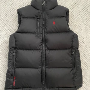 Svart dunväst från Polo Ralph Lauren - Svart dunväst från Polo Ralph Lauren med röd logga på bröstet. Västen har hög krage, tre dragkedjeförsedda fickor och quiltad design. Materialet är polyester med dunfyllning. Perfekt för lager-på-lager och chill stil. Pris kan självklart diskuteras.