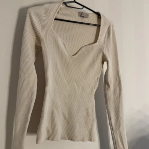 Ribbad vit långärmad topp - Snygg ribbad vit topp med lång ärm och djup v-ringning. Figurnära passform och stretchigt material som sitter skönt. Perfekt att styla med jeans eller kjol för en clean look.