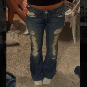 True Religion bootcut jeans blå, stl s - Säljer ett par blå bootcut jeans från True Religion med slitna detaljer och kontrastsömmar. Jeansen har låg midja, klassiska fickor med lock bak och tydliga slitningar framtill för en edgy look. Tillverkade i kraftig denim med snygg tvätt. 